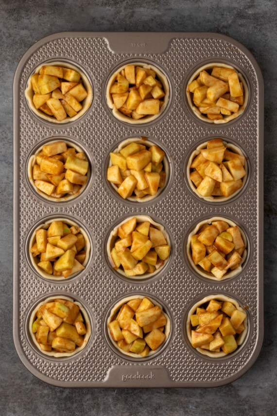 Mini pie crusts filled with apple pie filling inside a 12-well muffin tin.
