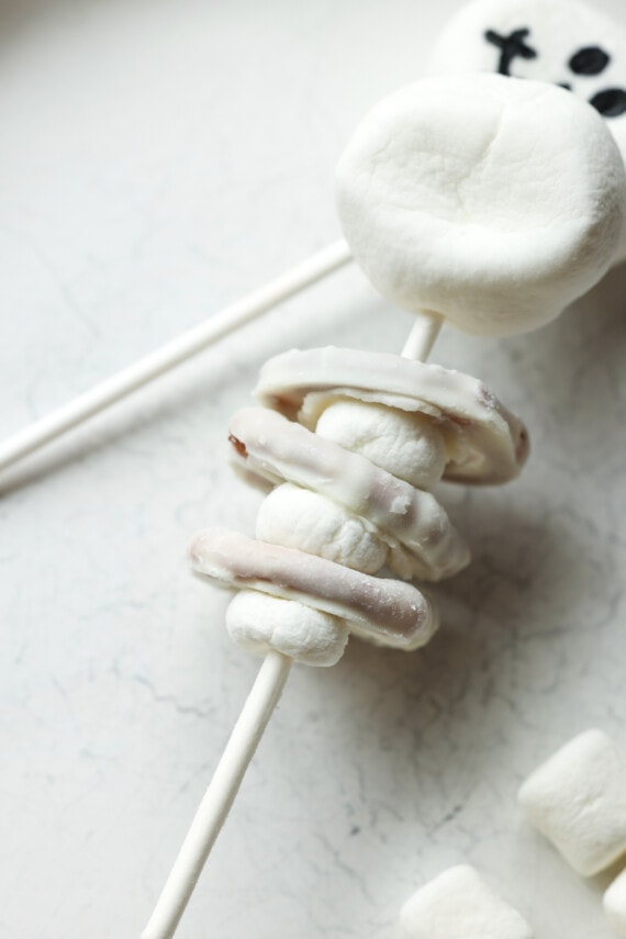 A lollipop stick threaded with mini marshmallows and white chocolate covered pretzels making edible skeleton decorations