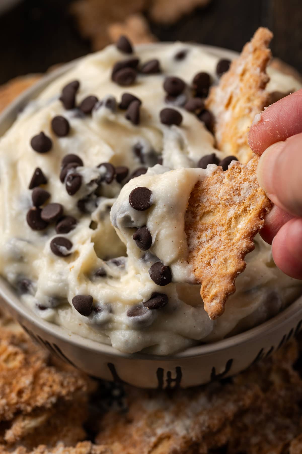A hand dips a cookie into a bowl of cannoli dip.