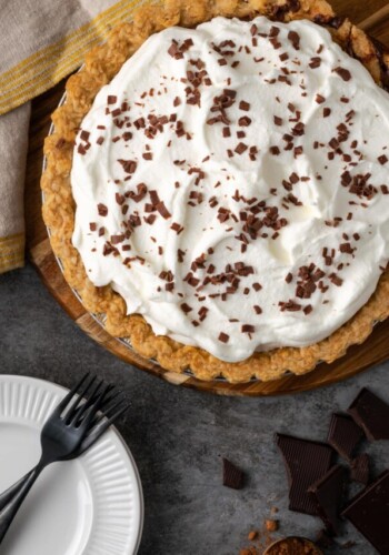 Overhead view of chocolate pudding pie topped with whipped cream and chocolate shavings, next to a fork on a white plate.