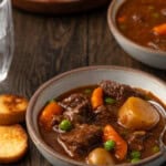 A bowl of Instant Pot beef stew on a wooden tabletop, with a plate of crostini in the background.