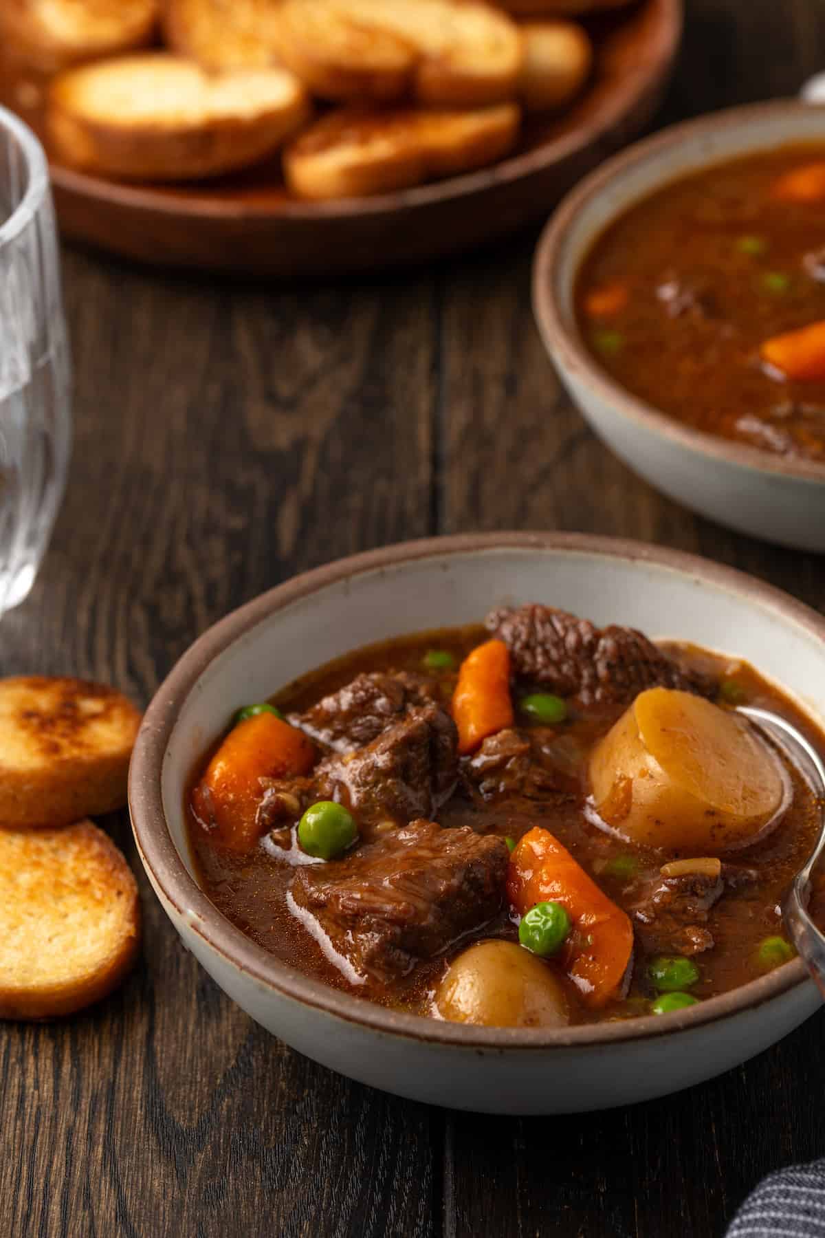 A bowl of Instant Pot beef stew on a wooden tabletop, with a plate of crostini in the background.