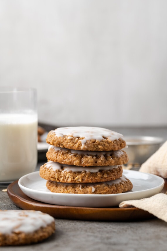 A stack of iced oatmeal cookies on a white plate with a glass of milk in the background.