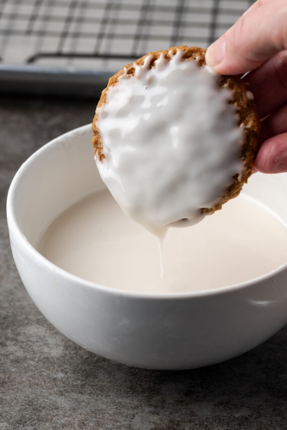 A hand holds an icing-covered oatmeal cookie above a bowl of vanilla icing.
