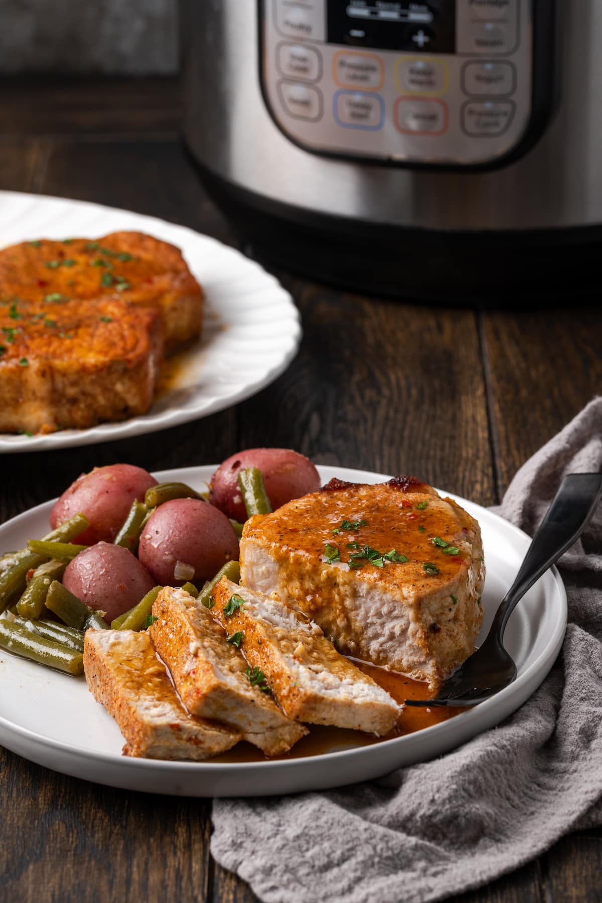 A pork chop cut into slices next to green beans and baby red potatoes on a white plate, with the instant pot in the background.