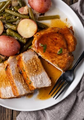 Overhead view of a pork chop cut into slices next to green beans and baby red potatoes on a white plate.
