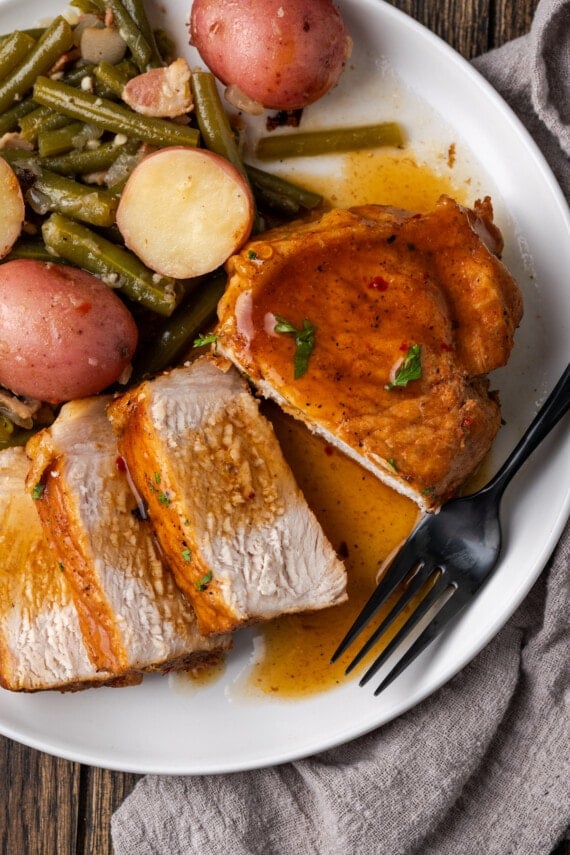 Overhead view of a pork chop cut into slices next to green beans and baby red potatoes on a white plate.