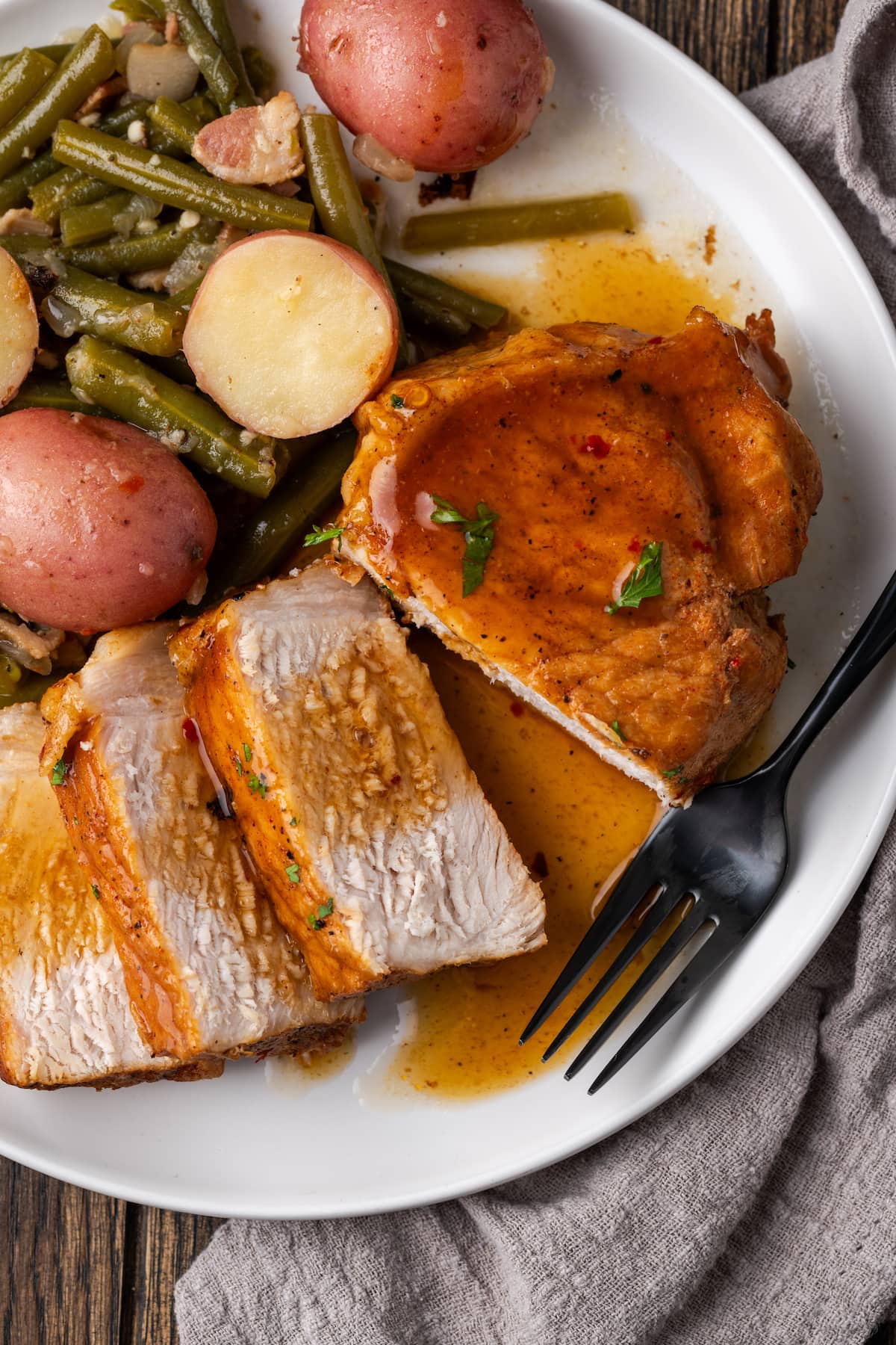Overhead view of a pork chop cut into slices next to green beans and baby red potatoes on a white plate.