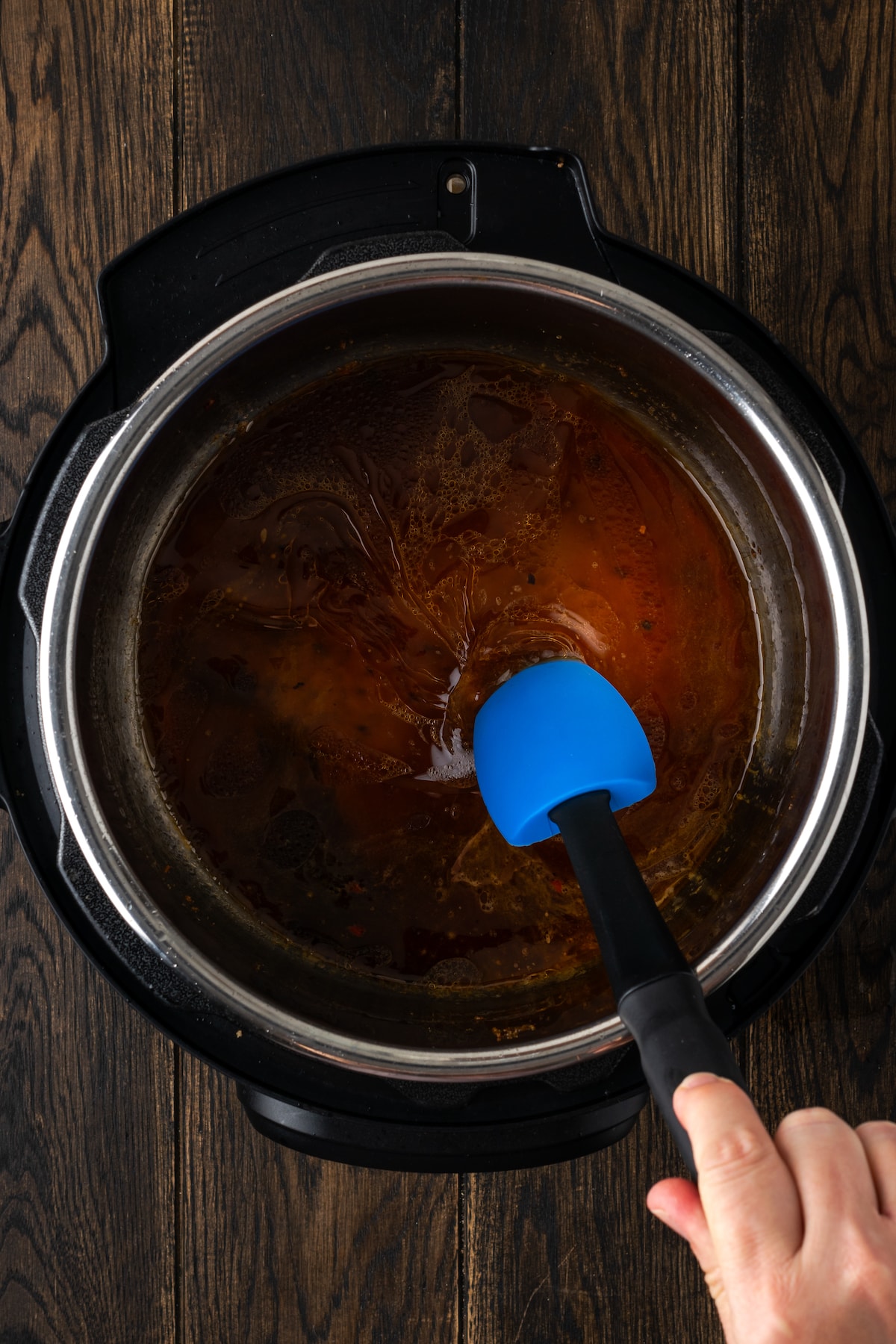 A hand uses a rubber spatula to deglaze the bottom of the instant pot.