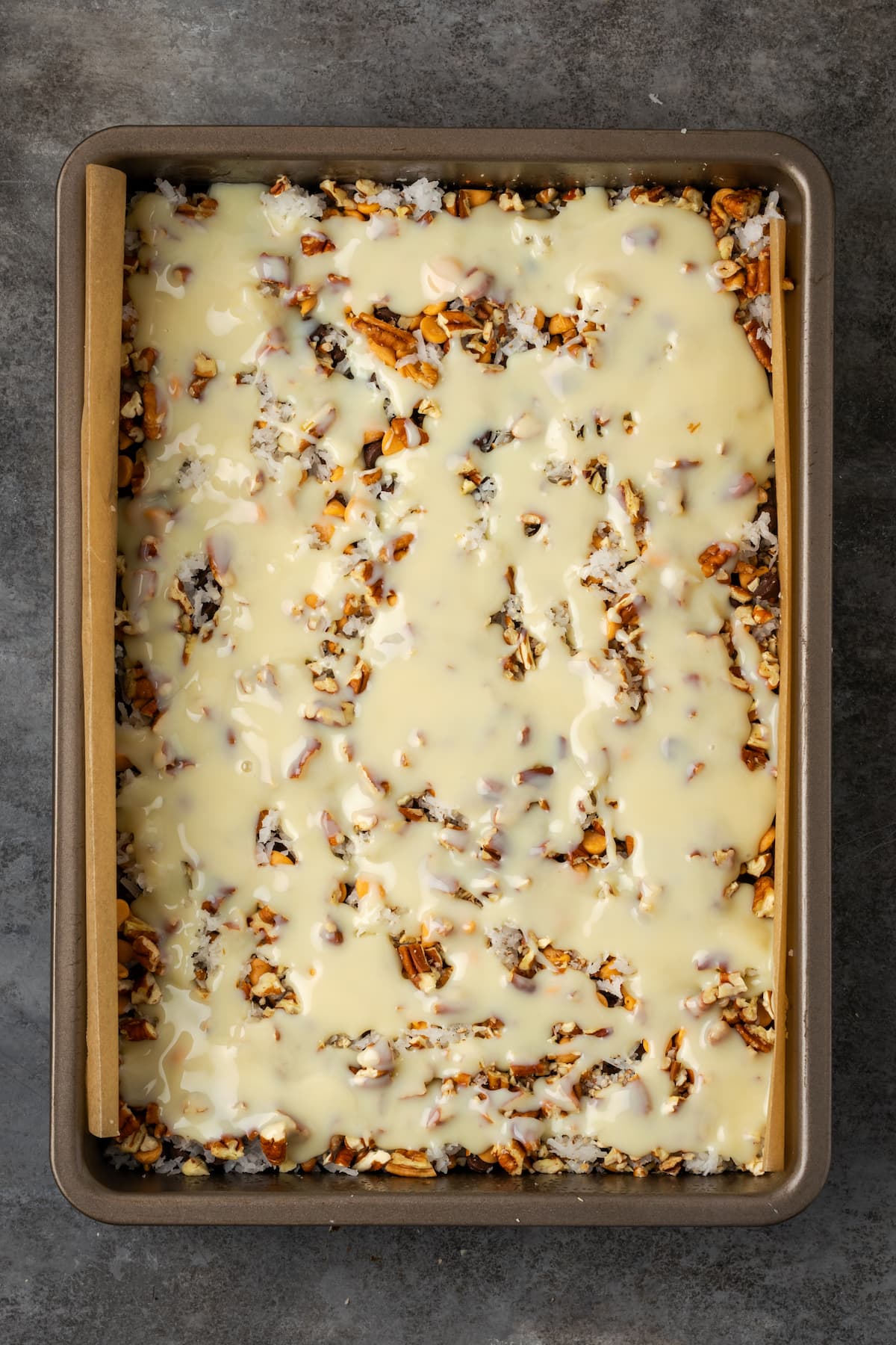 Magic bar ingredients covered with sweetened condensed milk in a lined baking pan.