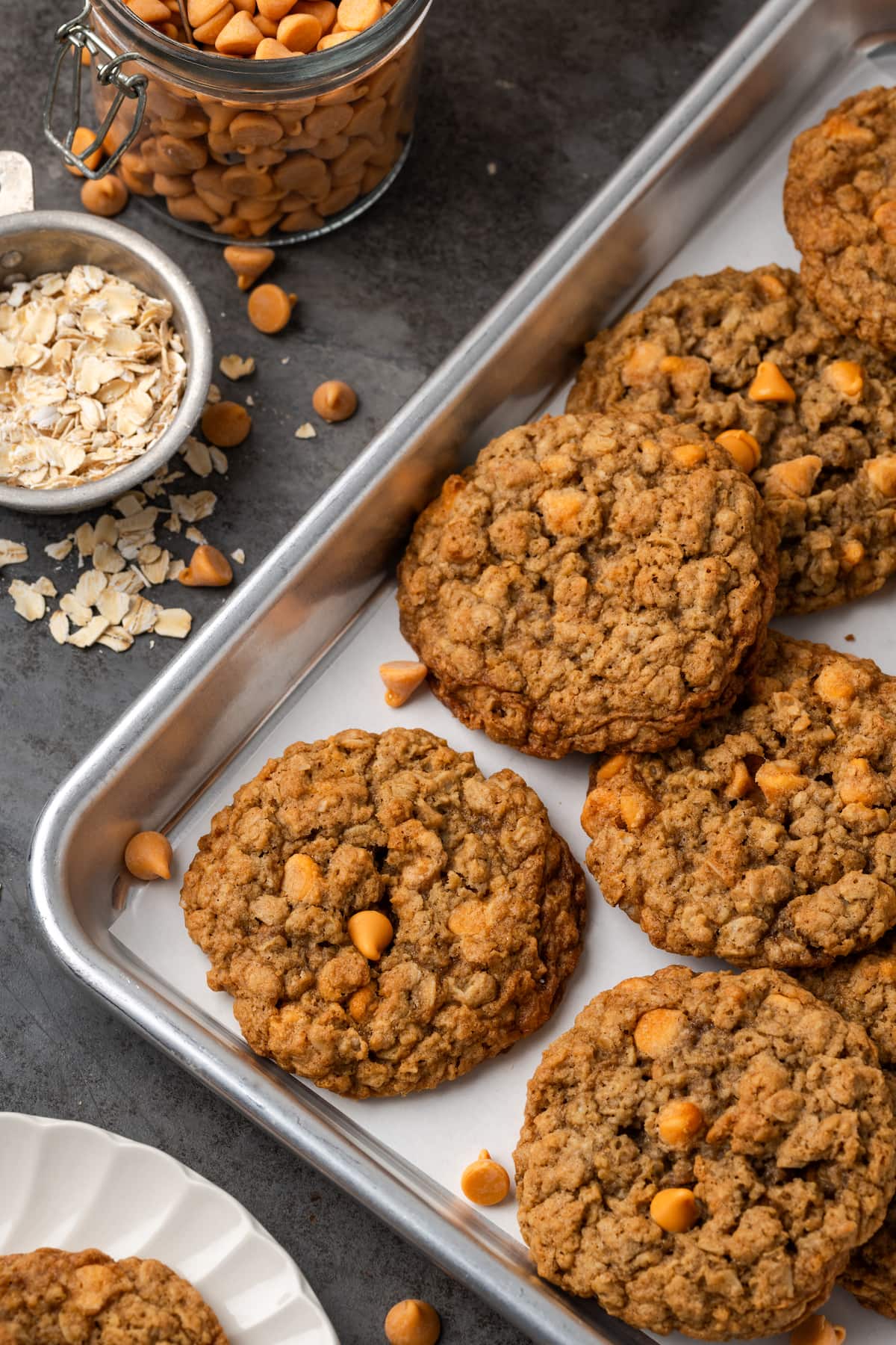 Assorted oatmeal butterscotch cookies on a baking sheet, next to small bowls of oats and butterscotch chips.