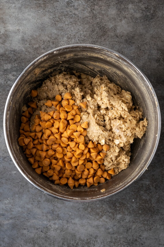 Butterscotch chips added to oatmeal cookie dough in a metal mixing bowl.