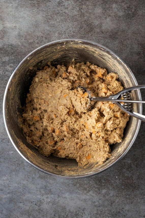 Oatmeal butterscotch cookie dough in a metal mixing bowl, with a cookie scoop.