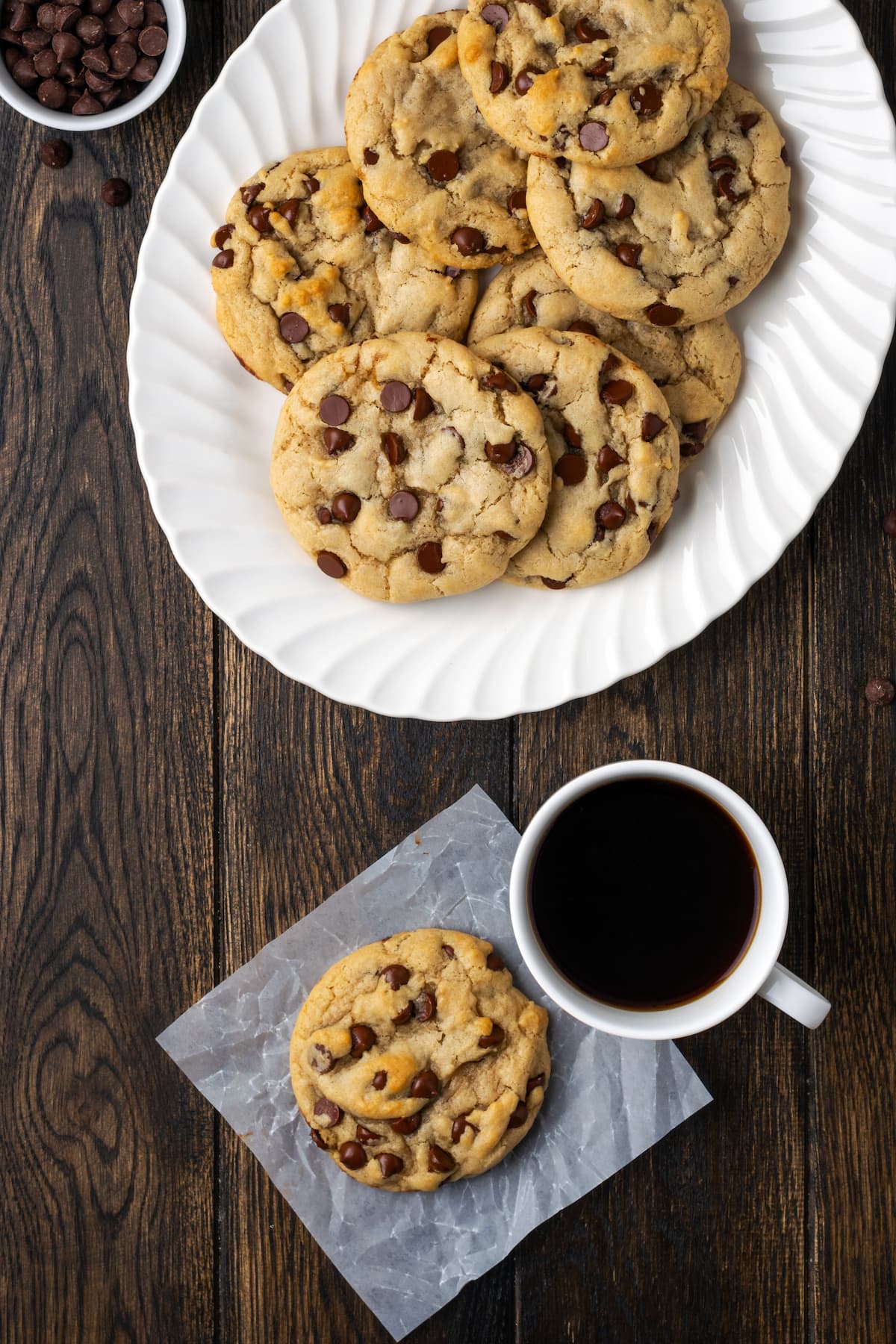 Overhead view of a chocolate chip Subway cookie on a piece of parchment paper next to a cup of coffee and more cookies on a white plate.