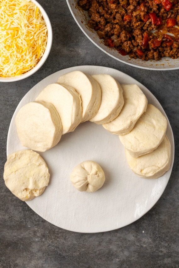 A filled biscuit dough ball on a plate next to more biscuit dough discs, and bowls of taco meat and shredded cheese.