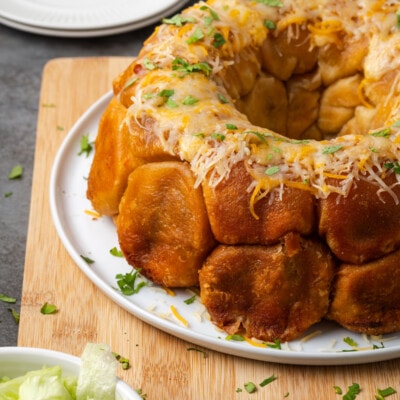Taco monkey bread topped with melted cheese on a white plate, next to a bowl of shredded lettuce.