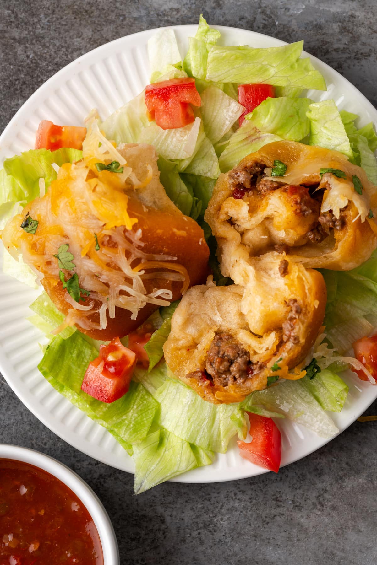 Overhead view of two taco monkey bread bites on top of shredded lettuce and diced tomatoes on a white plate, with one bread piece torn open to reveal the taco filling.