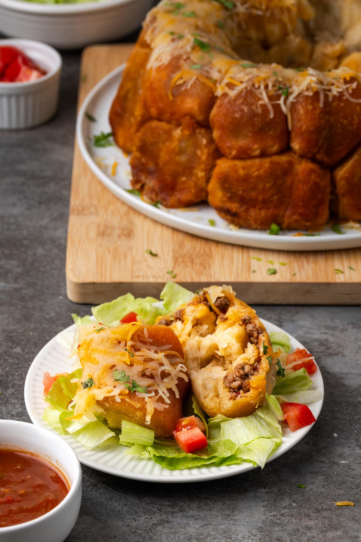 Taco monkey bread bites served on a plate with lettuce and diced tomatoes, with the full monkey bread in the background.