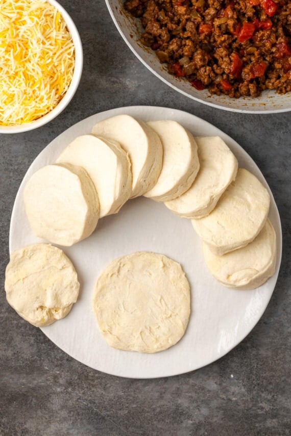 Biscuit dough flattened into a circle on a white plate next to more dough discs, and bowls of taco meat and shredded cheese.