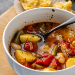 A bowl of instant pot vegetable soup with a spoon, with a cornbread muffin in the background.