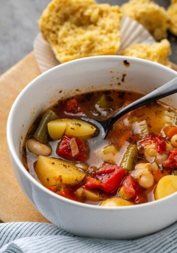 A bowl of instant pot vegetable soup with a spoon, with a cornbread muffin in the background.