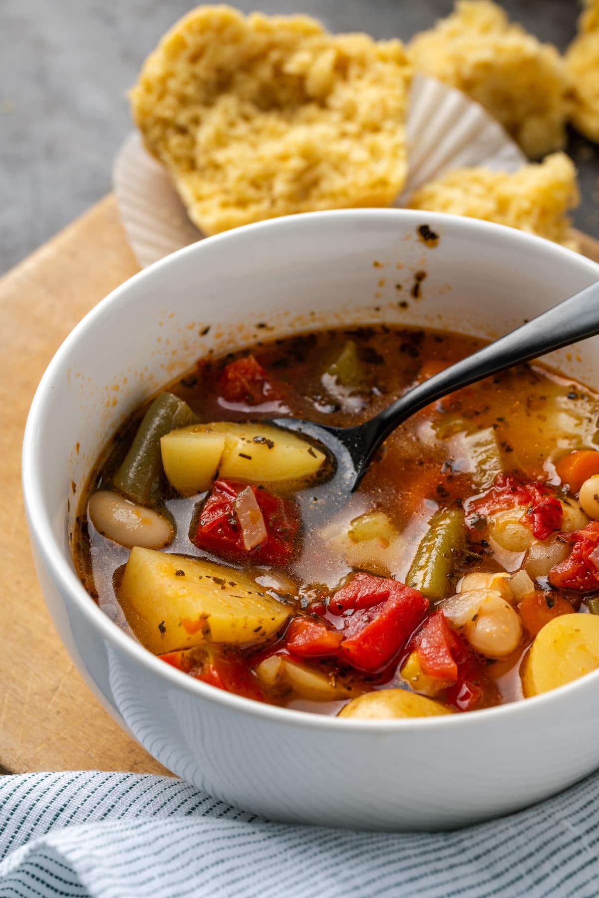 A bowl of instant pot vegetable soup with a spoon, with a cornbread muffin in the background.