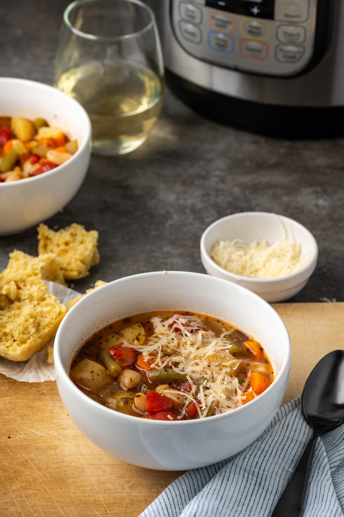 A bowl of vegetable soup on a wooden cutting board next to a cornbread muffin, with a second bowl of soup, a glass of white wine, and the instant pot in the background.