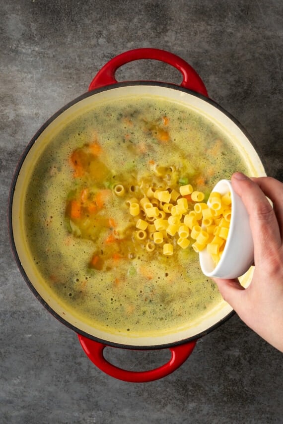A hand pours a cup full of pasta into a large pot of white bean soup.