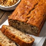 A homemade banana bread loaf with slices cut from the end, with a bowl of walnuts in the background.