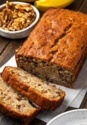 A homemade banana bread loaf with slices cut from the end, with a bowl of walnuts in the background.