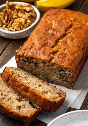 A homemade banana bread loaf with slices cut from the end, with a bowl of walnuts in the background.