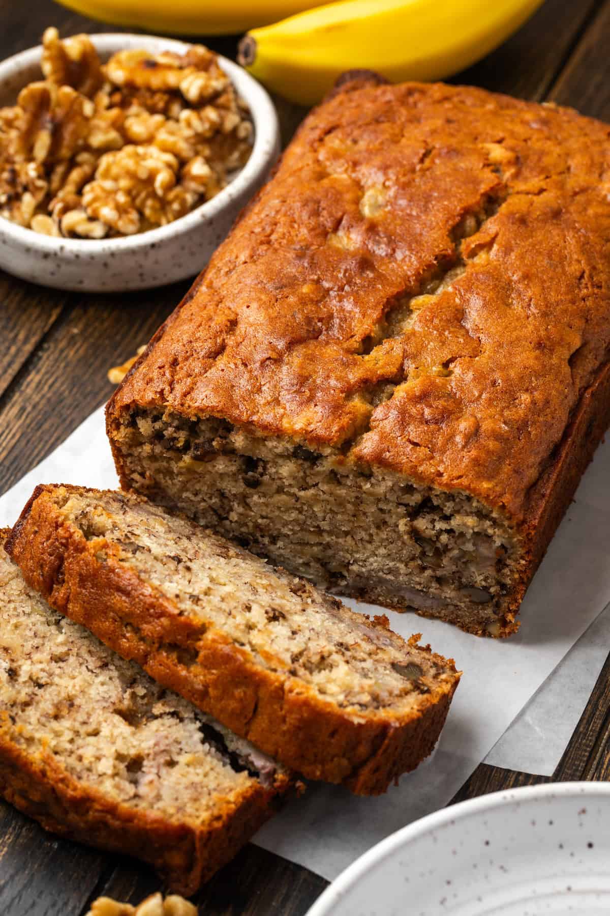 A homemade banana bread loaf with slices cut from the end, with a bowl of walnuts in the background.