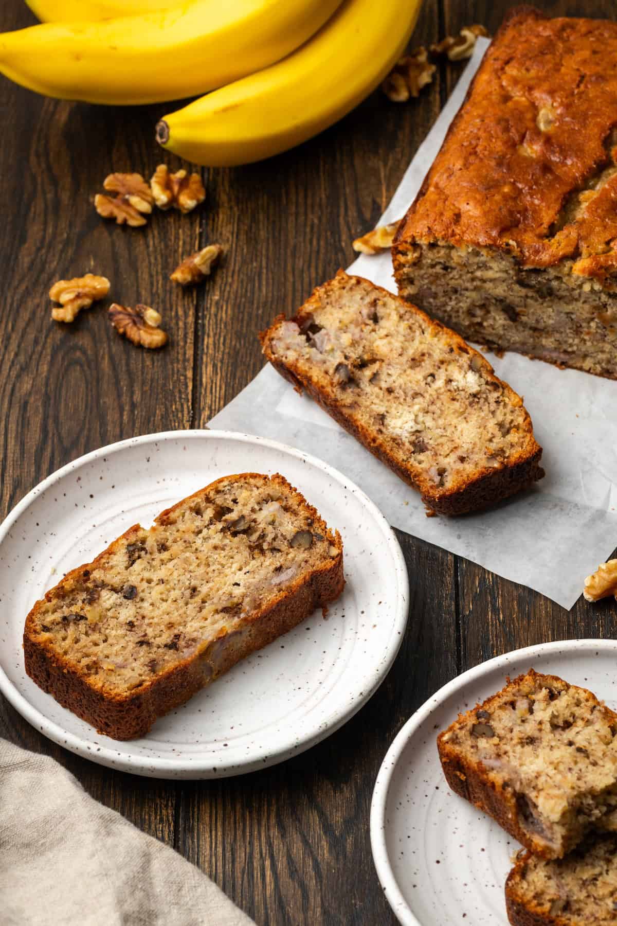 Banana bread slices on plates with the rest of the loaf next to bananas in the background.
