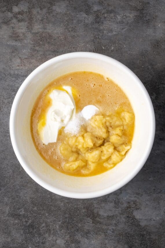 Mashed banana and sour cream added to the wet ingredients in a bowl.