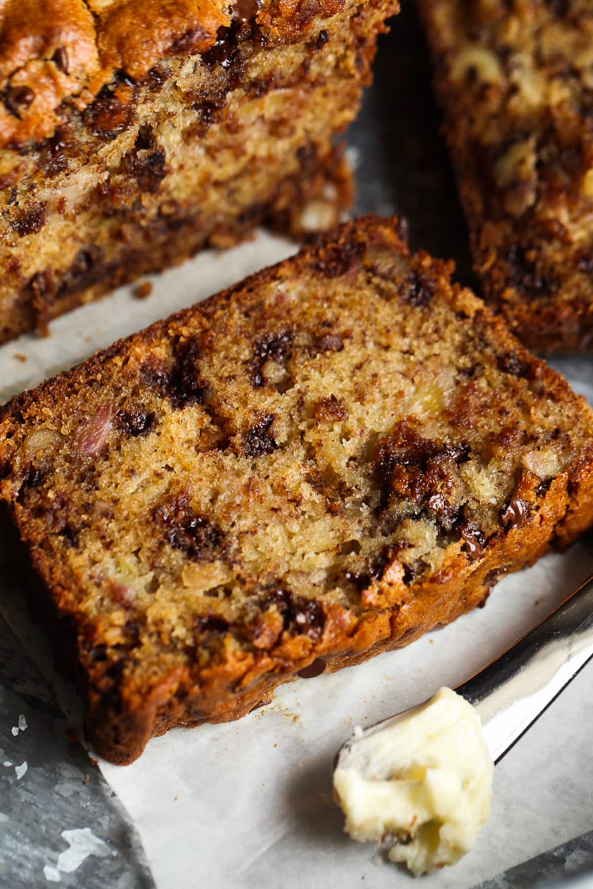 A slice of banana bread with chocolate chips and walnuts on a platter with a butter knife