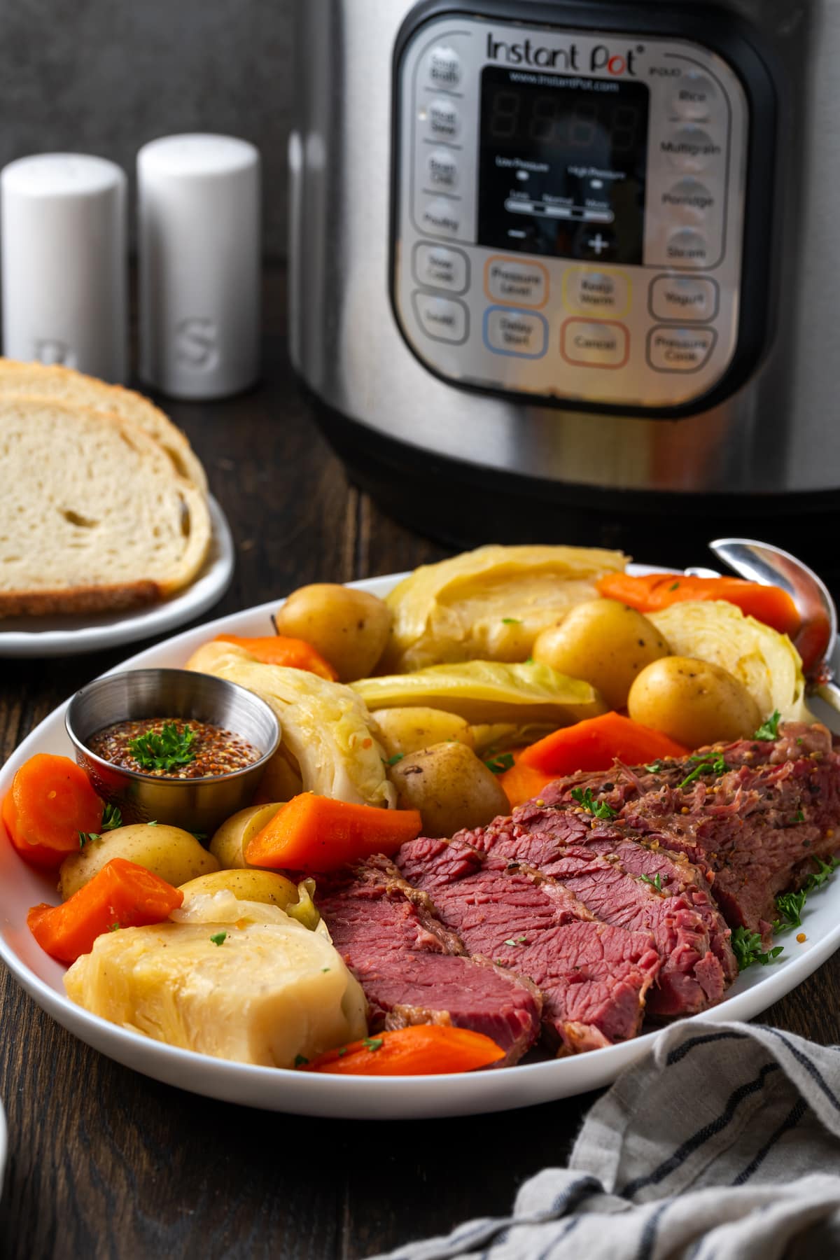 Sliced corned beef on a plate next to cabbage, potatoes, and carrots, with the Instant Pot and a place of bread slices in the background.