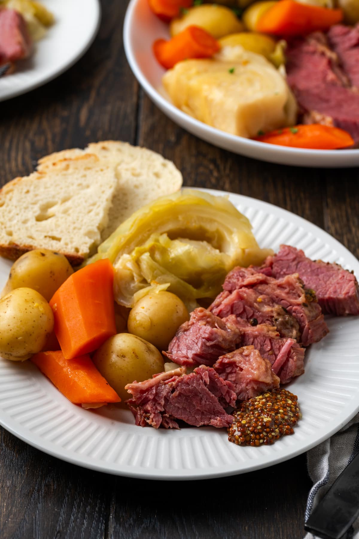 Sliced corned beef on a plate next to cabbage, potatoes, carrots, and bread slices, with a second plate in the background.