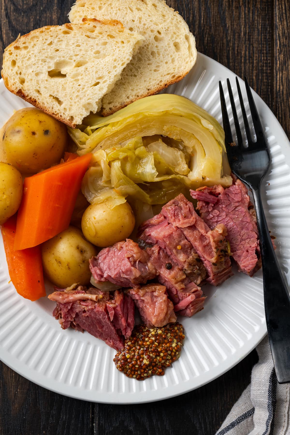 Overhead view of sliced corned beef on a plate with cabbage, potatoes, carrots, and bread slices, next to a fork.