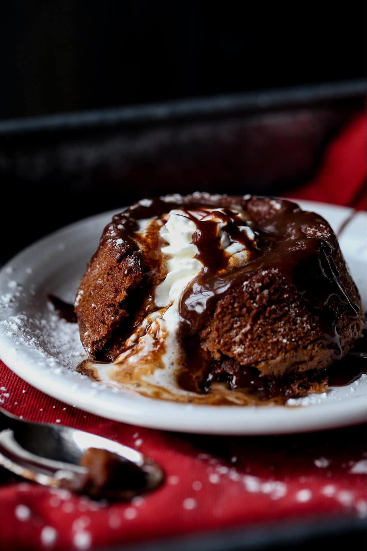 Chocolate Lava Cake topped with whipped cream and hot fudge melting from the inside on a white plate.