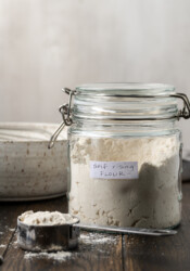 Side view of self rising flour in a mason jar, next to a measuring spoon with a bowl in the background.