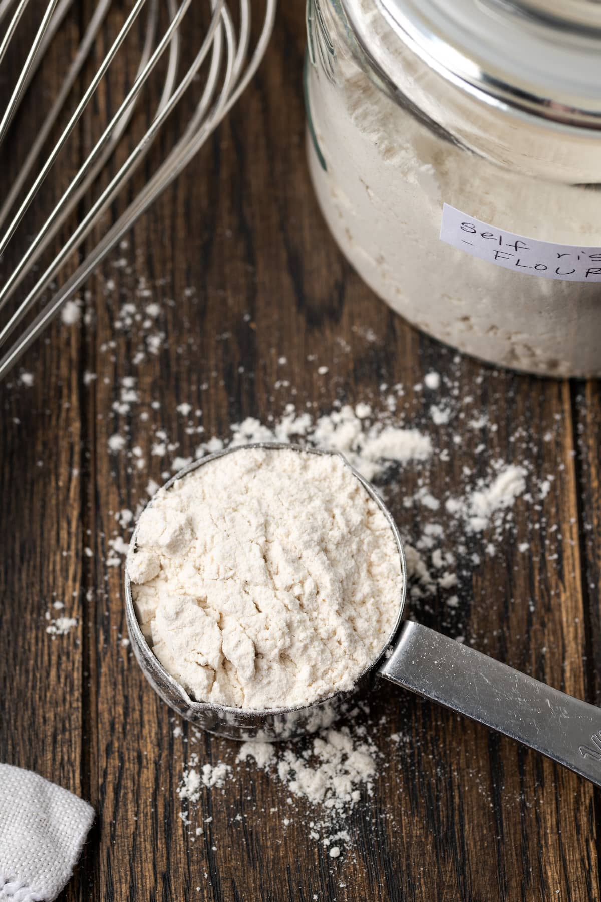 Overhead view of self rising flour in a measuring spoon next to more flour in a mason jar and a whisk.