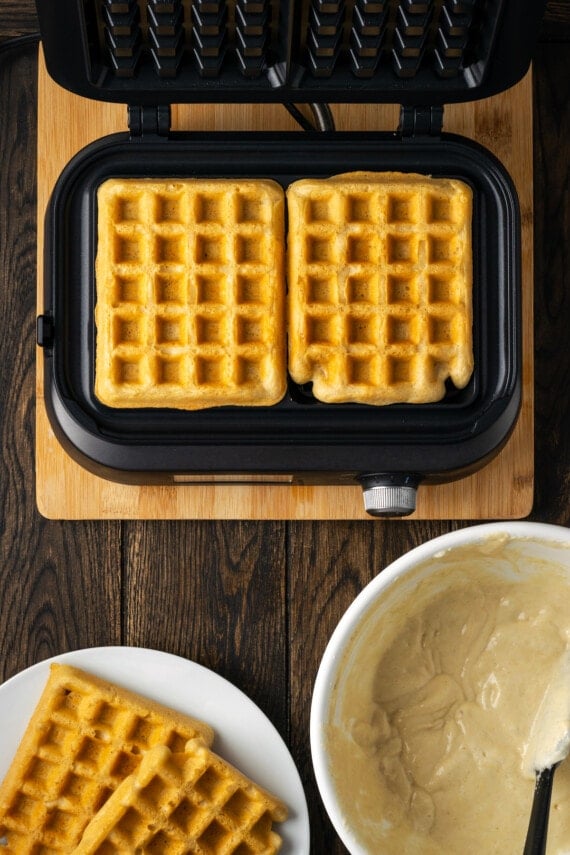 Two cooked waffles in a waffle iron, next to a bowl of waffle batter and a plate of finished waffles.