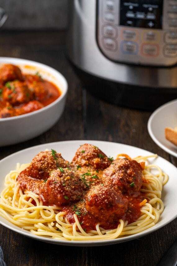 Instant pot meatballs and sauce served over a bed of spaghetti on a white plate, with the instant pot and a bowl of meatballs in the background.