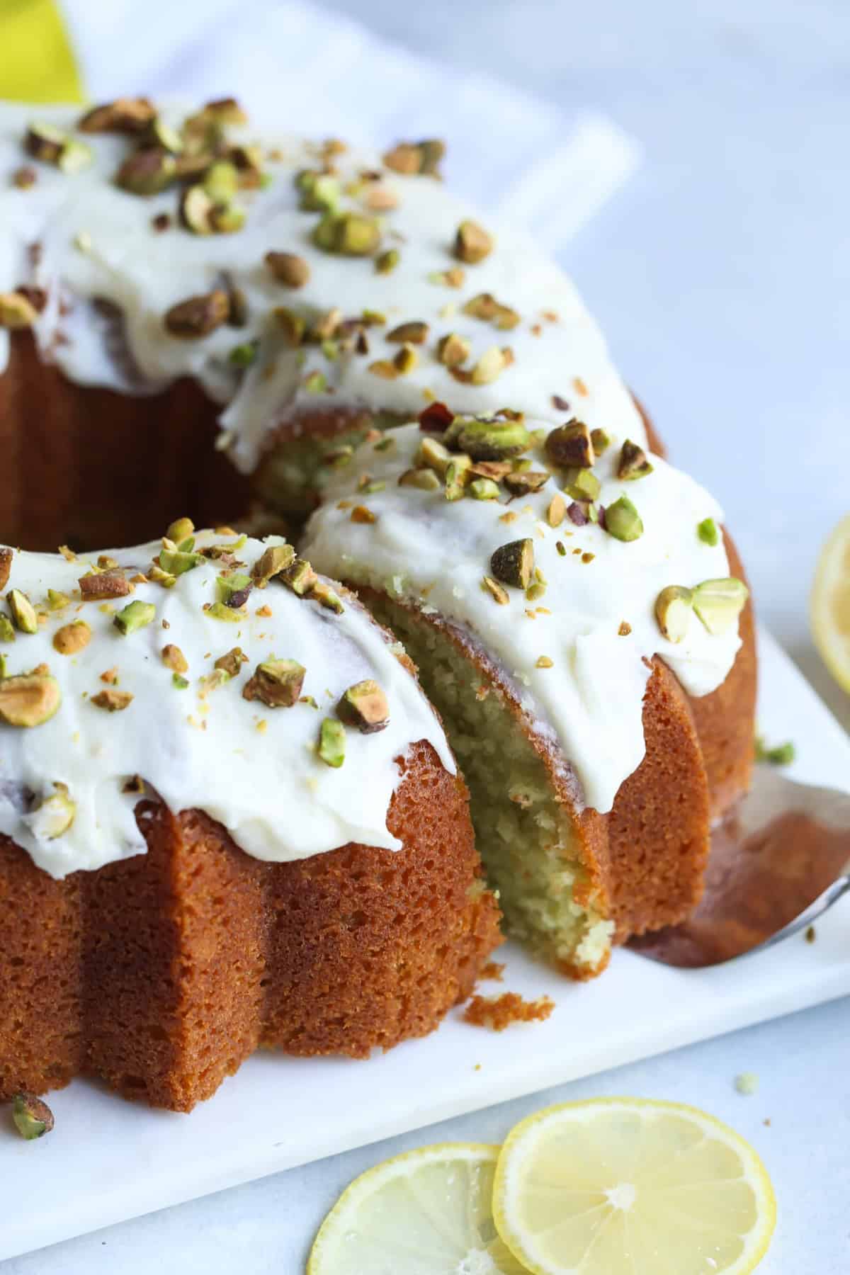 Frosted pistachio lemon bundt cake garnished with crushed pistachios on a square plate with a slice being served.