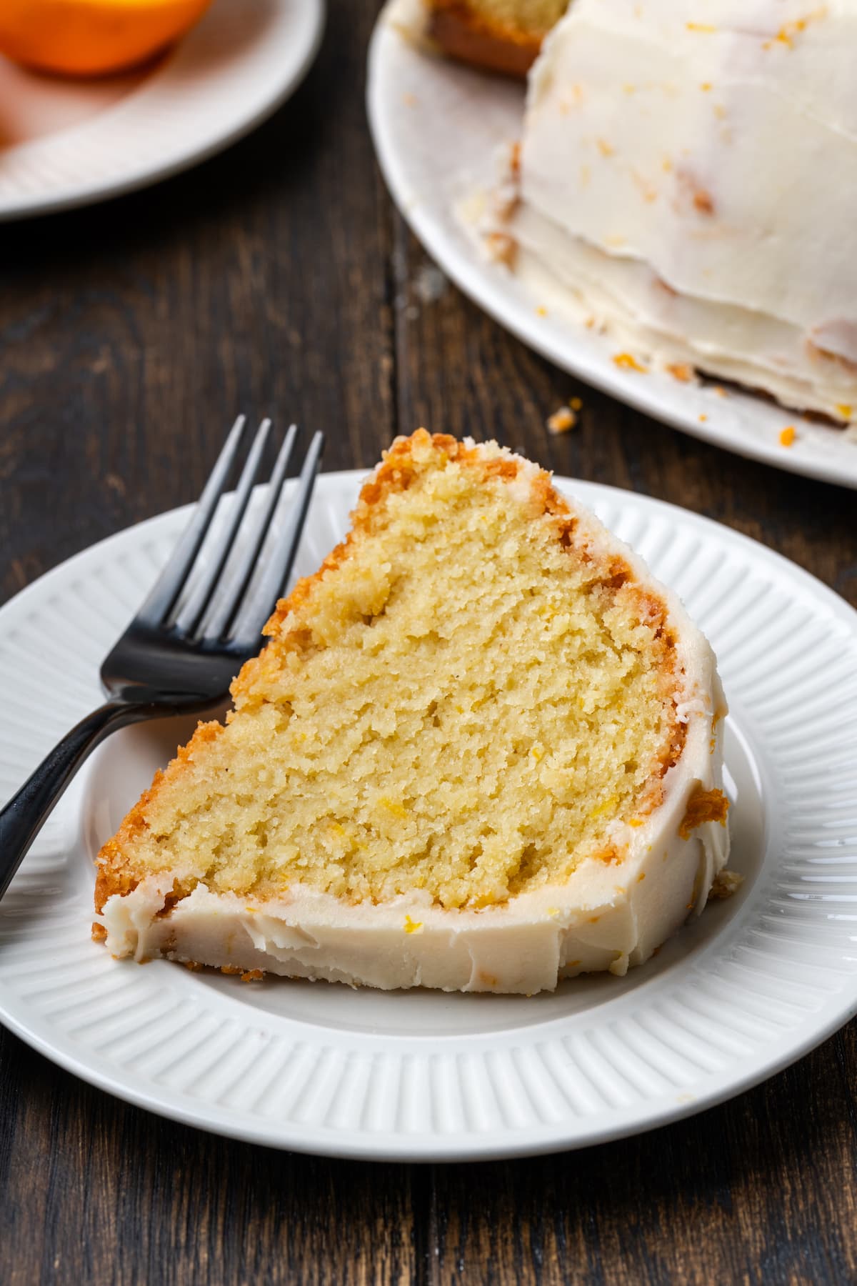 A slice of Meyer lemon bundt cake on a white plate next to a fork, with a corner of the rest of the cake in the background.