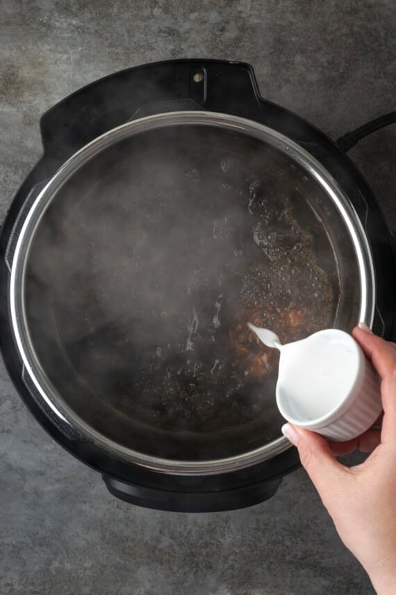 This juicy Instant Pot pork loin is smothered with a savory spice rub and out-of-this-world tender. Sear and cook your pork all in one pot! A hand pouring a cornstarch slurry from a small bowl into the cooking liquid inside the instant pot.