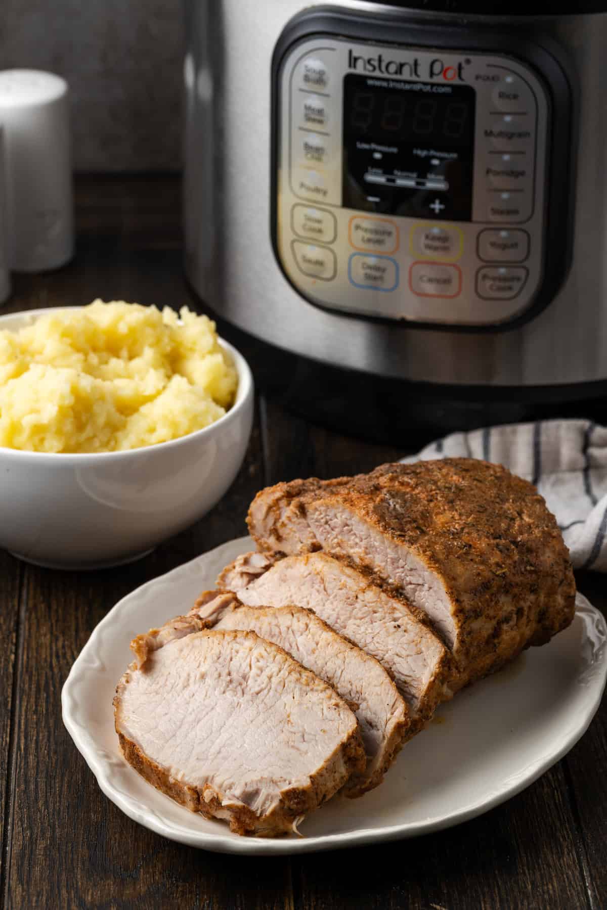 This juicy Instant Pot pork loin is smothered with a savory spice rub and out-of-this-world tender. Sear and cook your pork all in one pot! Pork loin sliced on a white platter, with a bowl of mashed potatoes and the instant pot in the background.