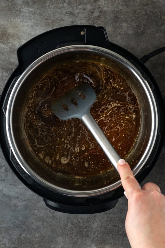 This juicy Instant Pot pork loin is smothered with a savory spice rub and out-of-this-world tender. Sear and cook your pork all in one pot! A hand using a spatula to deglaze the bottom of the instant pot with chicken broth.