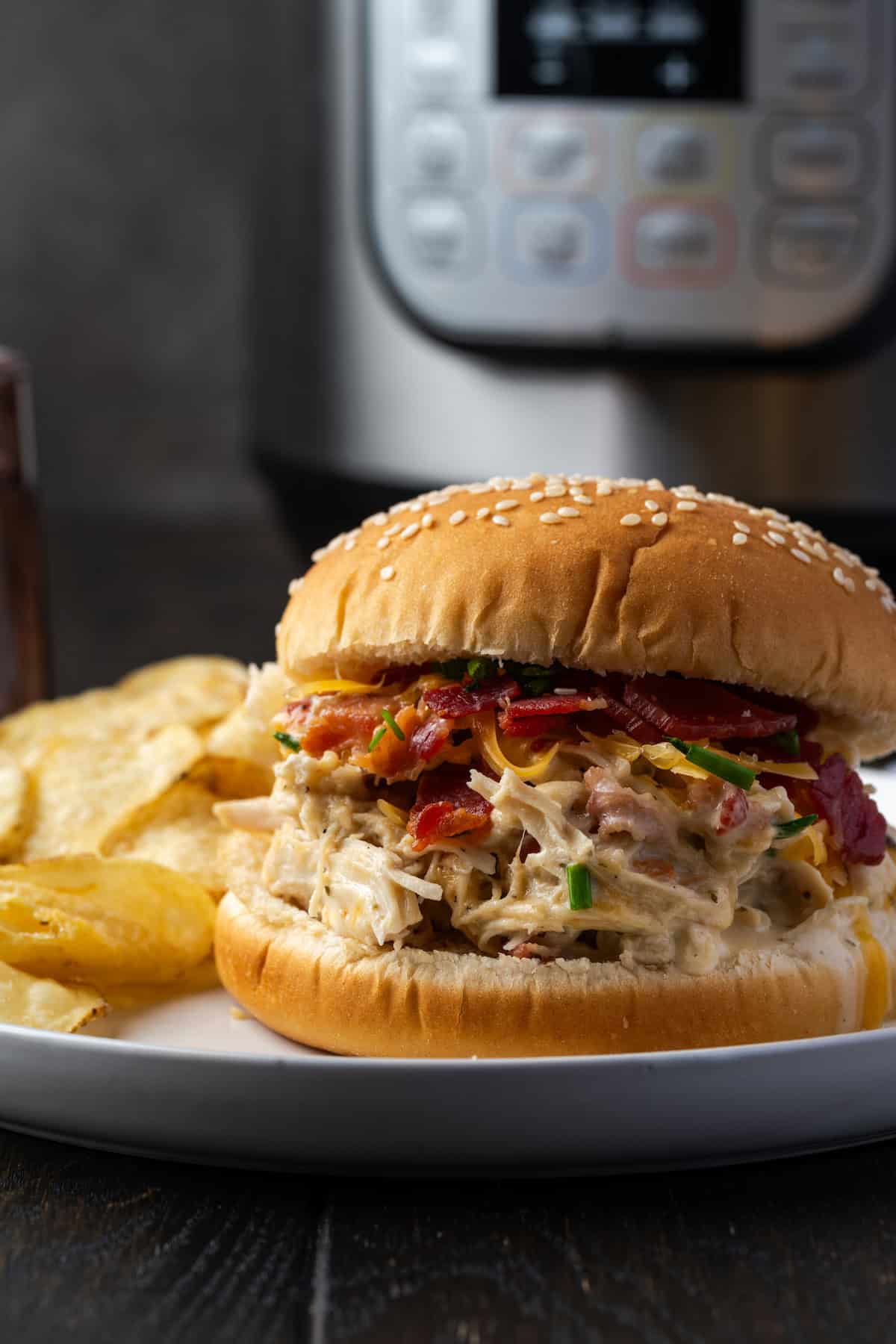 A crack chicken sandwich on a plate next to potato chips, with the Instant Pot in the background.