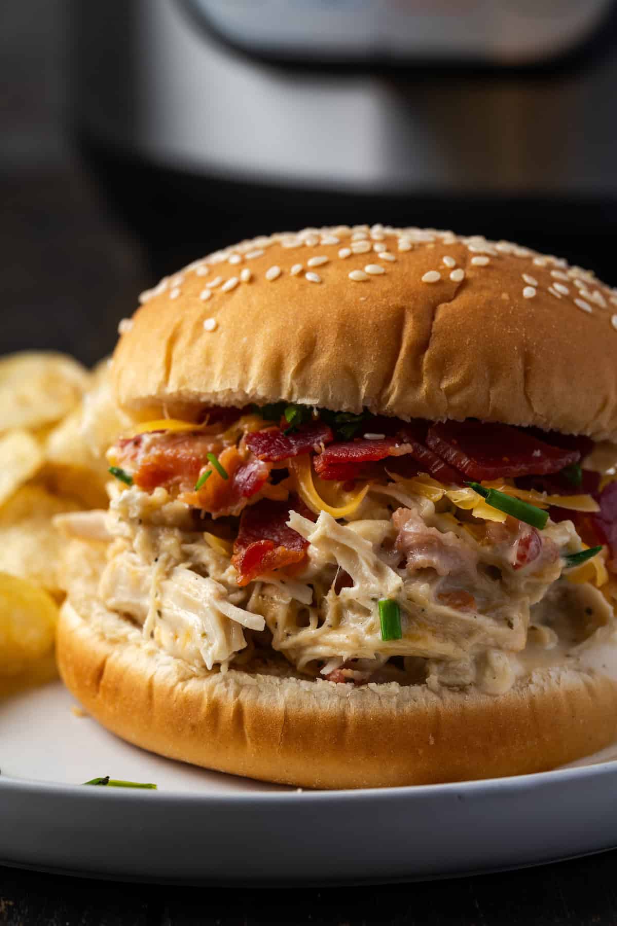 Close up of a crack chicken sandwich on a plate next to potato chips, with the Instant Pot in the background.